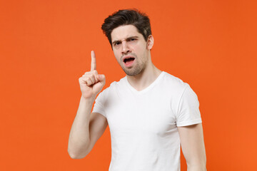 Fototapeta premium Angry dissatisfied young man wearing basic casual empty blank white t-shirt standing pointing index finger up screaming swearing isolated on bright orange colour wall background, studio portrait.