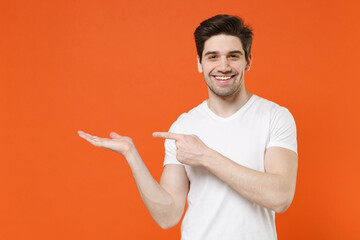 Smiling handsome young man 20s in basic casual empty blank white t-shirt standing pointing index finger hand aside on mock up copy space isolated on bright orange colour background, studio portrait.