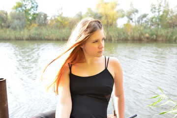 A blonde woman in a black long dress on a wooden pier on the river bank. Beautiful landscapes, green leaves and trees on the shore