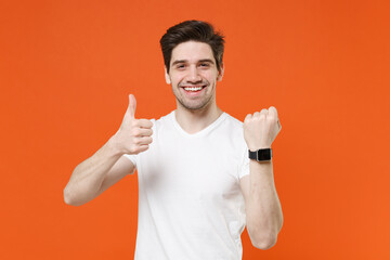 Smiling cheerful attractive handsome young man 20s in basic casual empty blank white t-shirt standing wearing smart watch showing thumb up isolated on bright orange colour background studio portrait.