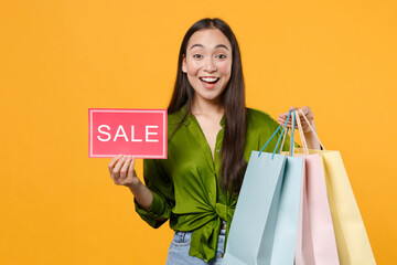 Excited young brunette asian woman 20s in basic green shirt standing hold package bags with purchases after shopping sign with SALE title looking camera isolated on yellow background, studio portrait.