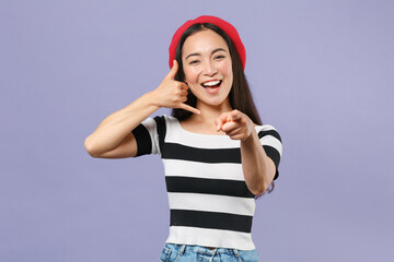 Obraz na płótnie Canvas Funny young asian woman in striped t-shirt red beret standing doing phone gesture like says call me back pointing index finger on camera isolated on pastel violet colour background studio portrait.