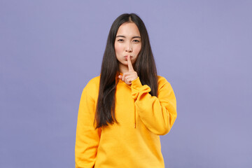 Secret young brunette asian woman wearing basic yellow hoodie standing saying hush be quiet with finger on lips shhh gesture looking camera isolated on pastel violet colour background studio portrait.