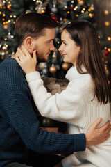 Close up side profile shot of adorable and tender couple looking each other and hugging. Christmas tree on background.