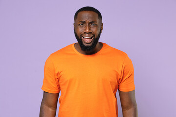 Shocked worried nervous young african american man 20s wearing basic casual orange t-shirt standing keeping mouth open looking camera isolated on pastel violet colour background, studio portrait.