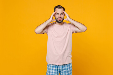 Naklejka premium Dissatisfied sick tired young bearded man in pajamas home wear put hands on head while resting at home isolated on bright yellow colour background studio portrait. Relax good mood lifestyle concept.