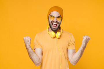 Overjoyed screaming young bearded man 20s wearing basic casual t-shirt headphones eyeglasses hat standing clenching fists doing winner gesture isolated on bright yellow background, studio portrait.