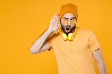 Shocked curious young bearded man in casual t-shirt headphones eyeglasses hat standing try to hear...