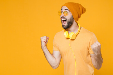 Overjoyed shocked screaming young bearded man 20s in basic casual t-shirt headphones eyeglasses hat standing clenching fists doing winner gesture isolated on bright yellow background, studio portrait.