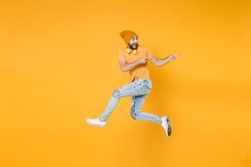 Fototapeta na wymiar Full length side view of shocked young man wearing basic casual t-shirt headphones hat jumping pointing index fingers aside on mock up copy space isolated on bright yellow background, studio portrait.
