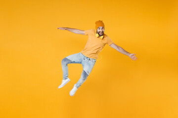 Fototapeta na wymiar Full length of excited cheerful surprised young man 20s wearing basic casual t-shirt headphones hat jumping spreading hands looking camera isolated on bright yellow colour background, studio portrait.
