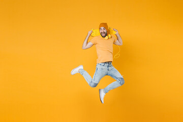 Full length of excited cheerful funny young man 20s wearing basic casual t-shirt headphones hat jumping hold skateboard looking camera isolated on bright yellow colour background, studio portrait. © ViDi Studio