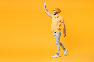 Fototapeta na wymiar Full length side view of cheerful young man wearing basic casual t-shirt headphones hat waving and greeting with hand as notices someone looking aside isolated on yellow background, studio portrait.