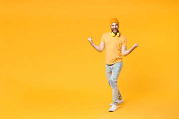 Fototapeta na wymiar Full length of overjoyed screaming young man in basic casual t-shirt headphones hat standing clenching fists doing winner gesture looking camera isolated on bright yellow background, studio portrait.