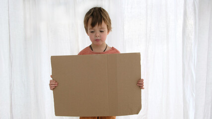 Upset little boy in orange holds large cardboard sheet with space for template standing near white curtain in light room