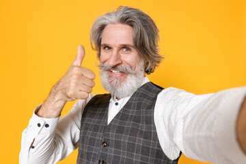 Close up of smiling elderly gray-haired business man in checkered waistcoat shirt doing selfie shot on mobile phone showing thumb up isolated on yellow background. Achievement career wealth concept.