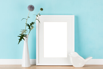 Fototapeta na wymiar Elegant vertical white picture frame with matte poster artwork mockup template for online shop with globe thistle in vase in front of pastel blue wall. Blank image area isolated with clipping path.
