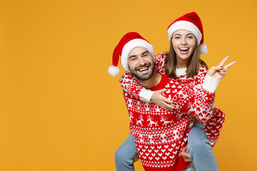 Laughing young Santa couple friends man woman in sweater Christmas hat giving piggyback ride to joyful, sit on back showing victory sign isolated on yellow background. Happy New Year holiday concept.