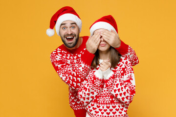 Surprised amazed young Santa couple friends man woman 20s in red sweater Christmas hat covering eyes with hands isolated on yellow background studio. Happy New Year celebration merry holiday concept.