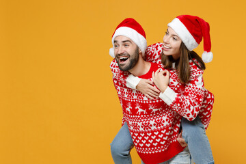 Cheerful funny young Santa couple friends man woman in sweater Christmas hat giving piggyback ride to joyful, sit on back looking aside isolated on yellow background. Happy New Year holiday concept.