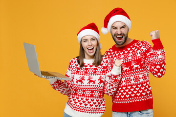 Happy young Santa couple friends man woman in sweater Christmas hat working on laptop pc computer doing winner gesture isolated on yellow background. Happy New Year celebration merry holiday concept.