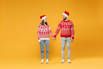 Full length of smiling young Santa couple friends man woman in sweater, Christmas hat hold hands looking at each other isolated on yellow background. Happy New Year celebration merry holiday concept.