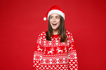 Surprised young brunette Santa woman in sweater, Christmas hat keeping mouth open looking aside isolated on red colour background, studio portrait. Happy New Year celebration merry holiday concept.