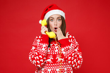 Shocked young Santa woman 20s wearing sweater Christmas hat talking on telephone covering mouth with hand isolated on red background studio portrait. Happy New Year celebration merry holiday concept.