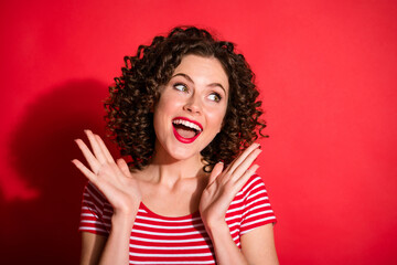 Photo of pretty cute curly young lady wear casual striped outfit looking empty space raising arms isolated red color background