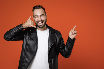 Laughing young bearded man in basic white t-shirt black leather jacket doing phone gesture like says call me back pointing index finger aside up isolated on orange colour background studio portrait.