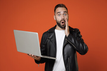 Shocked young bearded man 20s wearing basic white t-shirt black leather jacket standing working on laptop pc computer put hand on cheek isolated on bright orange colour background studio portrait.