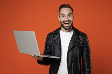 Excited funny young bearded man 20s wearing basic white t-shirt black leather jacket standing working on laptop pc computer looking camera isolated on bright orange colour background studio portrait.
