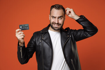 Naklejka premium Preoccupied young bearded man 20s wearing basic white t-shirt black leather jacket standing hold in hand credit bank card put hand on head isolated on bright orange colour background studio portrait.