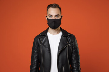 Young man 20s wearing basic white t-shirt black leather jacket face mask safe from coronavirus virus covid-19 during pandemic quarantine looking camera isolated on orange background studio portrait.