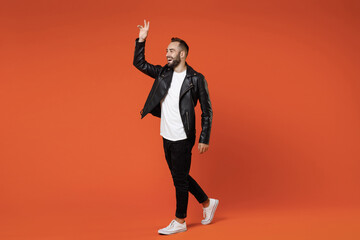 Fototapeta na wymiar Full length side view of cheerful young bearded man in basic white t-shirt black leather jacket standing waving and greeting with hand as notices someone isolated on orange background studio portrait.