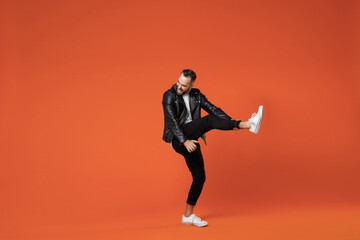Full length side view of crazy funny young bearded man in basic white t-shirt black leather jacket standing dancing rising leg like playing guitar isolated on orange colour background studio portrait.