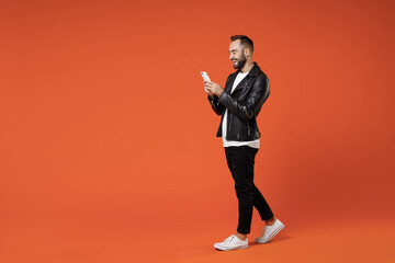 Fototapeta na wymiar Full length side view cheerful young bearded man in basic white t-shirt black leather jacket standing using mobile cell phone typing sms message isolated on orange colour background studio portrait.