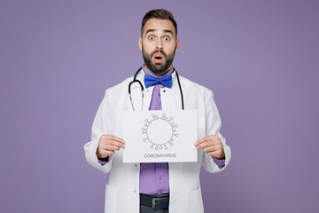 Shocked young bearded doctor man wearing white medical gown stethoscope hold nameplate coronavirus picture isolated on violet background studio portrait. Healthcare personnel health medicine concept.