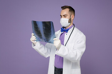 Young doctor man in medical gown face mask safe from coronavirus virus covid-19 hold X-ray of lungs fluorography roentgen isolated on violet background. Healthcare personnel health medicine concept.