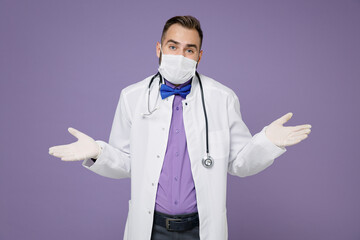 Confused young doctor man in medical gown stethoscope face mask to safe from coronavirus virus covid-19 spreading hands isolated on violet background. Healthcare personnel health medicine concept.