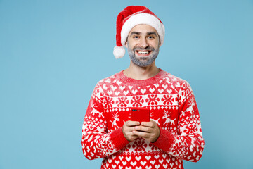 Smiling Northern bearded man frozen snow face in Santa hat Christmas sweater using mobile phone typing sms message isolated on blue background. Happy New Year celebration holiday winter time concept.
