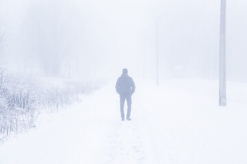 One young adult man in black clothes walking on sidewalk in heavy mist in white winter day. Foggy air. Poor visibility. Back view.