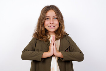 Beautiful little girl standing against white background,  keeps palms together, has pleased...