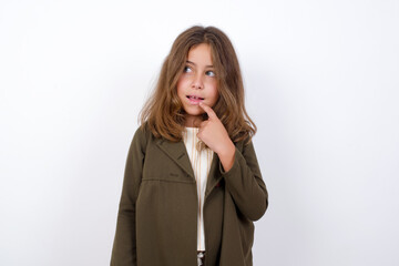 Beautiful little girl standing against white background,  with thoughtful expression, looks to the camera, keeps hand near face, bitting a finger thinks about something pleasant.