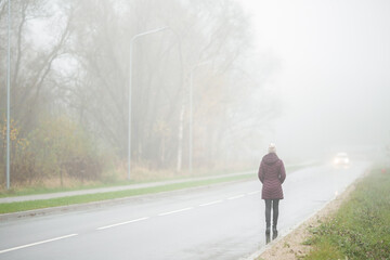 Young adult woman in dark clothes without reflective element walking unsafety on road in foggy day. Back view.