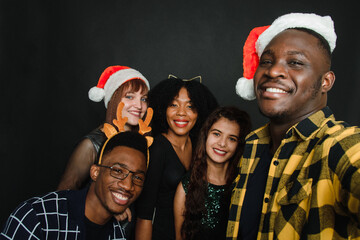 A group of friends of different nationalities take a Christmas selfie in the studio. Young men and women in santa hats and deer antlers are having fun together