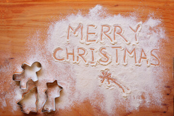Words 'Merry Christmas' written in spread flour and a variety of cookie cutters on a wooden chopboard.