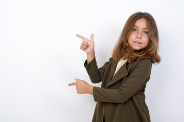 Beautiful little girl standing against white background, points aside with  surprised expression with mouth opened, shows something amazing. Advertisement concept.