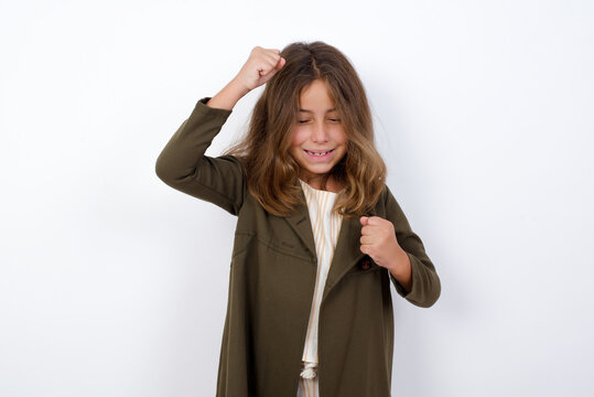 Attractive Beautiful Little Girl Standing Against White Background, Celebrating A Victory Punching The Air With His Fists And A Beaming Toothy Smile.