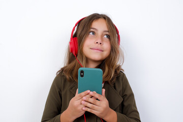 Happy Beautiful little girl standing against white background, listening to music with earphones using mobile phone.